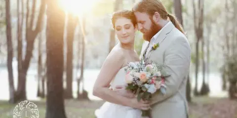 A bride and groom embracing in a wooded area near a river, with the bride holding a bouquet.