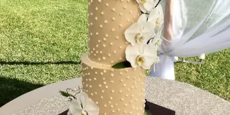 A two-tiered beige wedding cake decorated with white orchids and dried branches, sitting on a wooden stand outdoors.