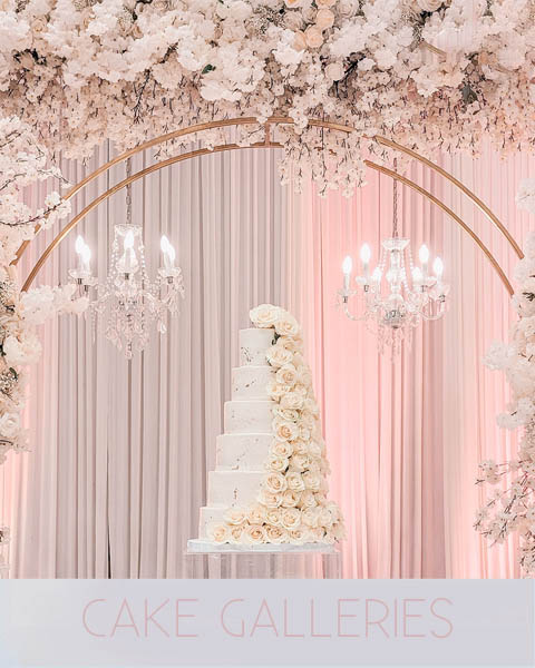 A white tiered wedding cake with cascading roses displayed under a floral arch and chandeliers.