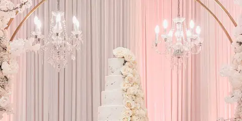 A white tiered wedding cake with cascading roses displayed under a floral arch and chandeliers.