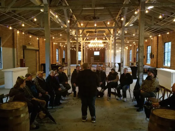 A group of people seated in a circle inside a rustic warehouse or distillery venue with wooden barrels.