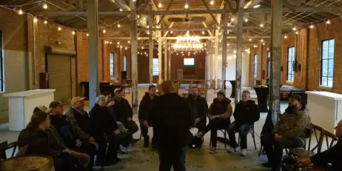 A group of people seated in a circle inside a rustic warehouse or distillery venue with wooden barrels.