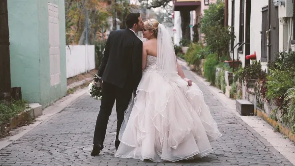 A bride and groom sharing a kiss on a cobblestone street, with the bride wearing a white gown and veil and the groom in a dark suit.