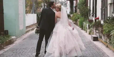 A bride and groom sharing a kiss on a cobblestone street, with the bride wearing a white gown and veil and the groom in a dark suit.