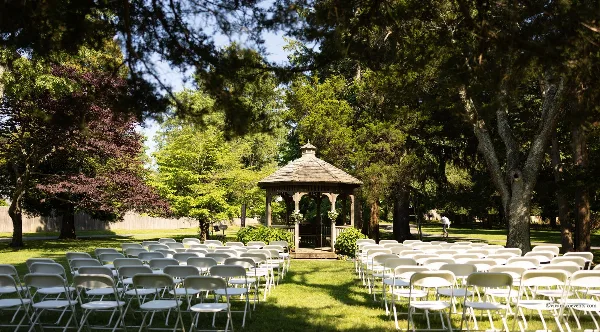 An outdoor wedding ceremony setup featuring rows of white chairs arranged on a lawn facing a wooden gazebo.