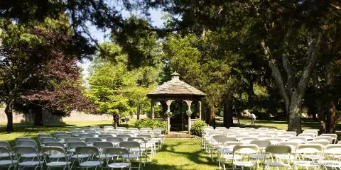 An outdoor wedding ceremony setup featuring rows of white chairs arranged on a lawn facing a wooden gazebo.