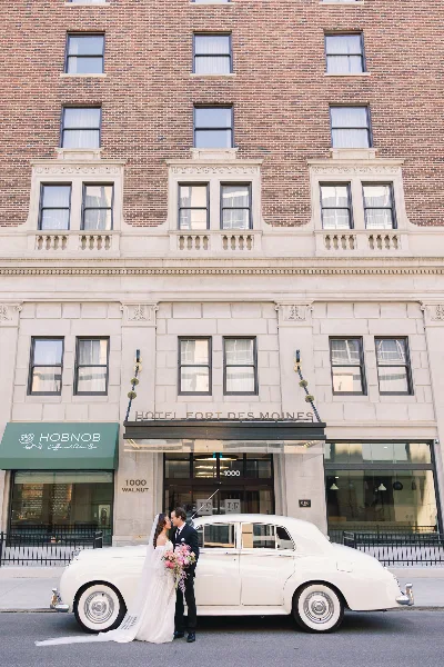 A bride and groom kissing in front of a white vintage car parked outside the Hotel Fort Des Moines.
