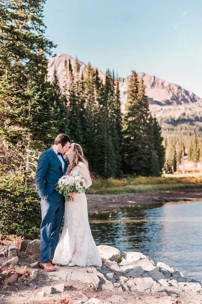 A bride and groom kissing on a rocky shore by a lake with pine trees and mountains in the background.