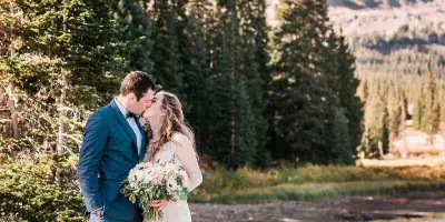 A bride and groom kissing on a rocky shore by a lake with pine trees and mountains in the background.