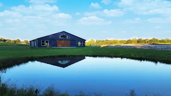 Aerial view of a dark blue barn-style wedding venue building on a grassy farm with a pond in the foreground.