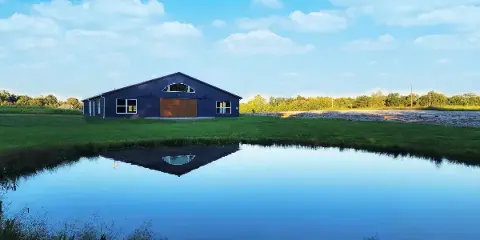 Aerial view of a dark blue barn-style wedding venue building on a grassy farm with a pond in the foreground.