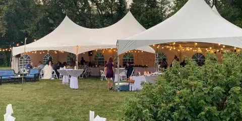 White event tent set up on a lawn with tables, chairs, and string lights for a wedding or party.