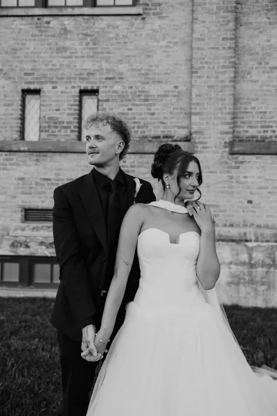 Black and white portrait of a bride and groom standing together in front of a brick wall.