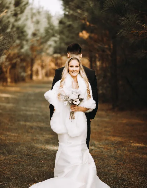 A bride and groom standing together in a wooded outdoor area, with the bride wearing a white gown and fur stole while holding a bouquet.