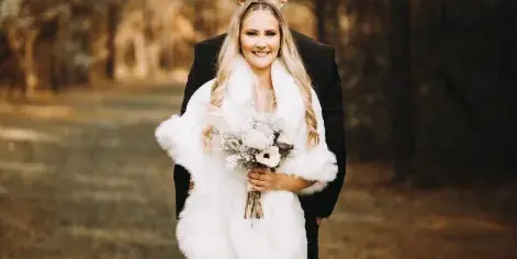 A bride and groom standing together in a wooded outdoor area, with the bride wearing a white gown and fur stole while holding a bouquet.