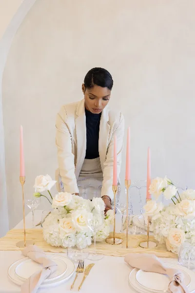 A professional event planner in a white blazer arranging white floral centerpieces and tall pink candles on a formal dining table.
