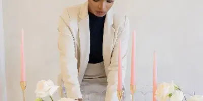 A professional event planner in a white blazer arranging white floral centerpieces and tall pink candles on a formal dining table.