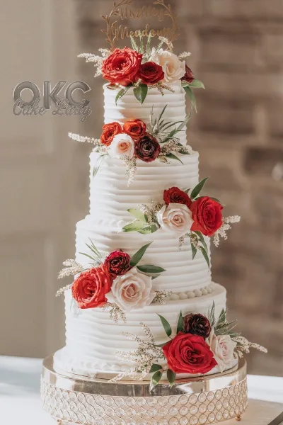 A four-tier white wedding cake decorated with red and cream roses, greenery, and a gold 'Mr & Mrs' topper.