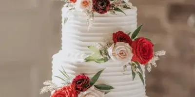 A four-tier white wedding cake decorated with red and cream roses, greenery, and a gold 'Mr & Mrs' topper.