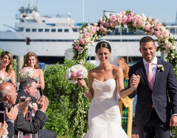 A bride and groom walking down the aisle during an outdoor wedding ceremony with a floral arch and a large ship in the background.