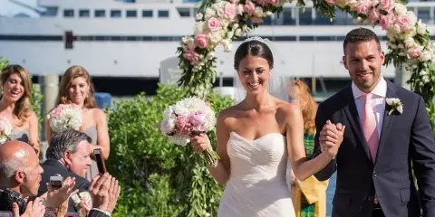 A bride and groom walking down the aisle during an outdoor wedding ceremony with a floral arch and a large ship in the background.