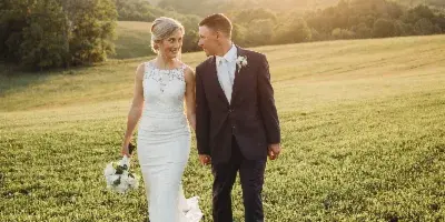 A bride and groom walking hand-in-hand across a grassy field during golden hour.