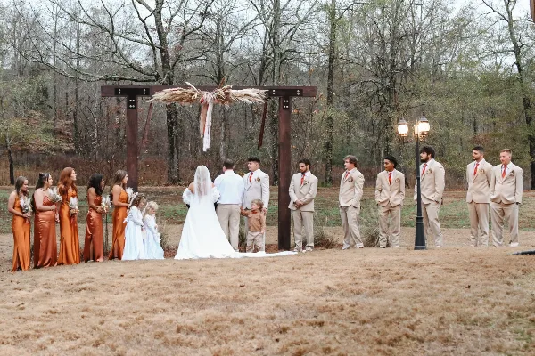 A wedding party stands at a wooden arch during an outdoor ceremony in a wooded area.