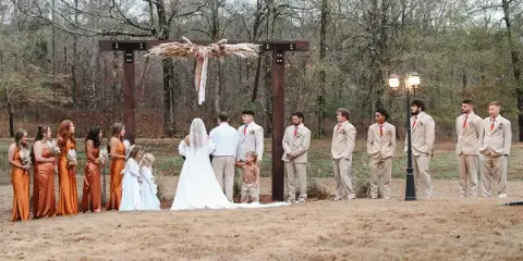 A wedding party stands at a wooden arch during an outdoor ceremony in a wooded area.