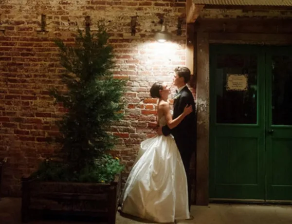 Bride and groom embracing in front of an exposed brick wall with a green door and potted plant.