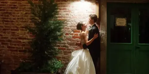 Bride and groom embracing in front of an exposed brick wall with a green door and potted plant.