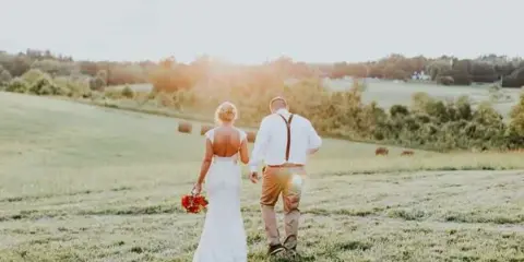A bride and groom walking away from the camera in a grassy field at sunset.
