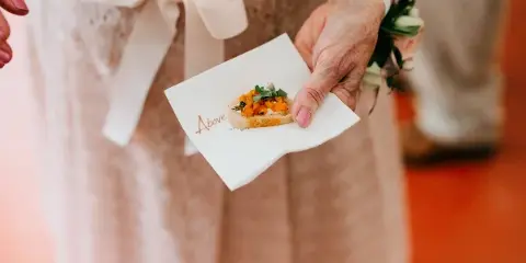 A person in a formal dress holds a small appetizer on a branded white napkin.