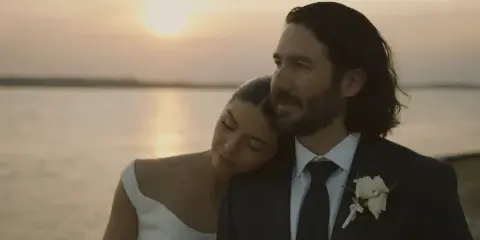 Cinematic still of a bride and groom embracing at sunset by the water