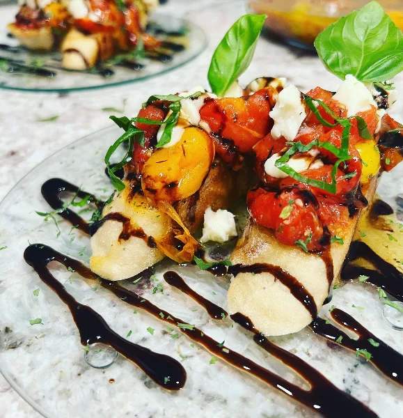 A close-up of a plated appetizer featuring bread topped with tomatoes, cheese, and basil, garnished with a balsamic drizzle.