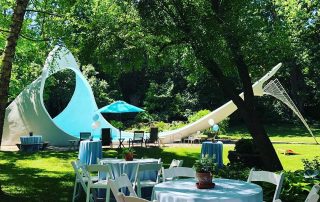 An outdoor event setup with white chairs, tables, and a large white tent in a grassy area.