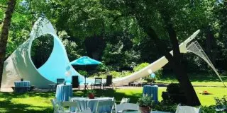 An outdoor event setup with white chairs, tables, and a large white tent in a grassy area.
