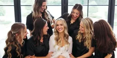 A bride in a white dress is surrounded by her bridesmaids in black dresses, all with styled hair, in a bright room.