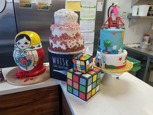 A display of custom cakes, including a tiered floral cake, a Rubik's cube cake, and a Russian nesting doll cake, with a Whisk Cake Creations box in the center.