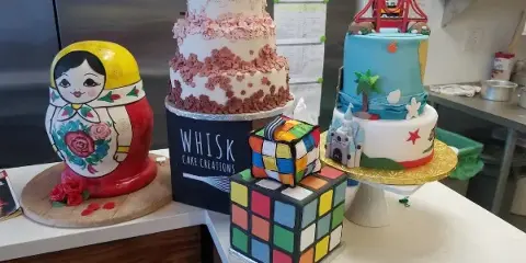 A display of custom cakes, including a tiered floral cake, a Rubik's cube cake, and a Russian nesting doll cake, with a Whisk Cake Creations box in the center.