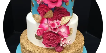 A three-tiered custom cake featuring a gold-dusted base, a white middle tier, and a blue top tier, decorated with vibrant pink and red fondant roses and gold leaves.