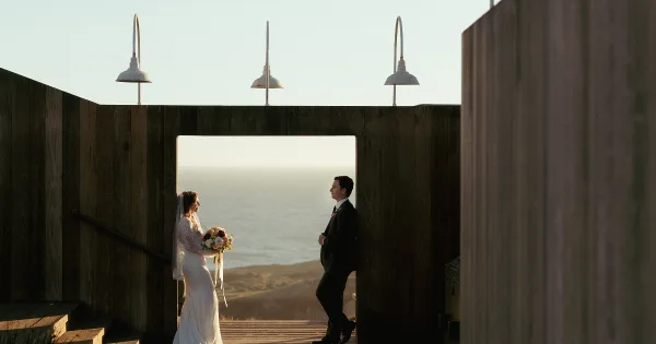 A bride and groom standing in a wooden doorway with an ocean view in the background.