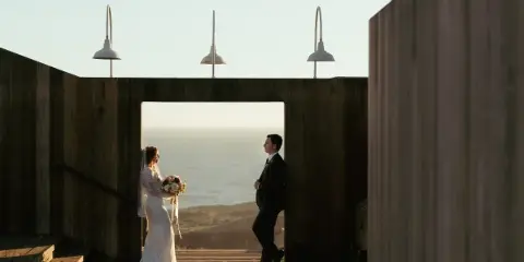 A bride and groom standing in a wooden doorway with an ocean view in the background.