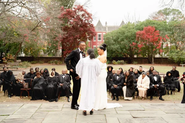 A bride and groom standing with an officiant during an outdoor wedding ceremony with seated guests.
