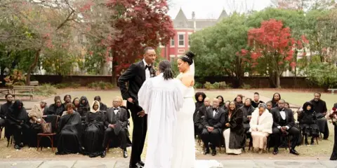 A bride and groom standing with an officiant during an outdoor wedding ceremony with seated guests.