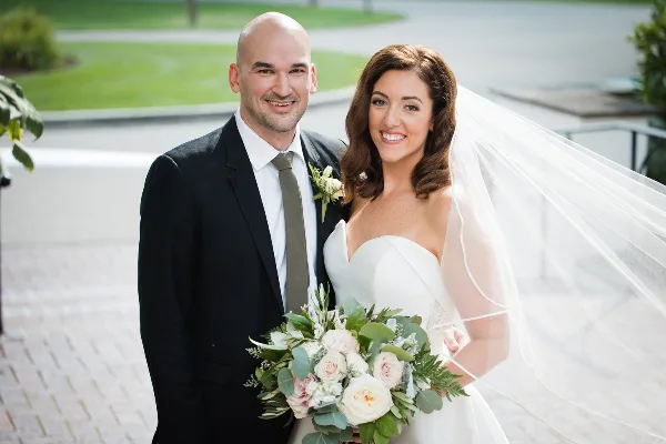 A smiling bride and groom standing together outdoors, with the bride holding a bouquet of white and pink roses.