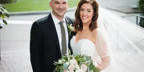 A smiling bride and groom standing together outdoors, with the bride holding a bouquet of white and pink roses.