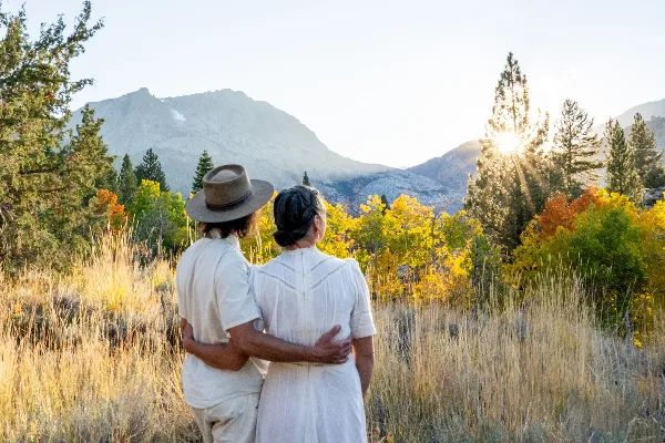A couple standing in a grassy field with a mountain range and sunset in the background.