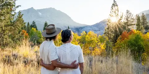 A couple standing in a grassy field with a mountain range and sunset in the background.