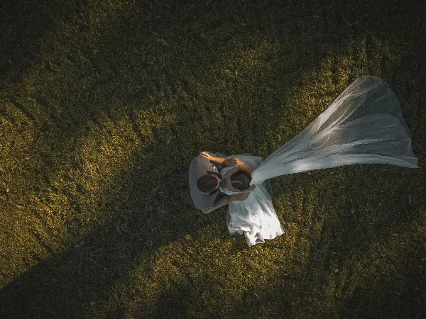 Aerial view of a bride and groom embracing on a grassy field, with the bride's veil flowing in the wind.