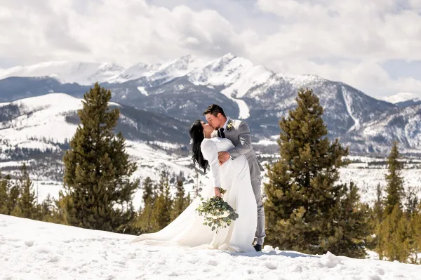 A bride and groom sharing a kiss on a snowy mountain slope with evergreen trees and snow-capped peaks in the background.